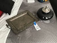 Photo showing the metal tray with floral embossed border and handles, and the black vanity jar with silver-tone lid on a textured surface.