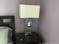 Full view of the table lamp on a dark wooden nightstand showing the cream rectangular shade and clear glass base.