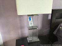 Tall modern table lamp with clear ribbed rectangular glass base and white fabric shade, slightly wider than the base, on a dark wooden table against a purple wall.