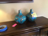 Pair of turquoise vases with paisley designs shown from a side angle on a wooden surface.