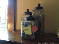 Three decorative glass canisters with dark lids grouped on a wooden mantel near a window.