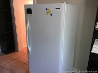 White upright freezer closed, showing full front and side view with handle and tape tag attached.