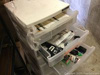 Tall white multi-drawer plastic cabinet with three drawers open showing various bike repair items inside under basement lighting.