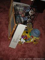 Photo showing black metal dog crate, wooden pet gate, blue pet bowl, pet care bottles, dog boots, harness, dog toys scattered on maroon carpet.