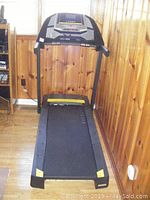 Full view of treadmill standing against wood-paneled wall, plugged in