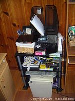 Front view of black four-tier shelf loaded with office supplies including shredder, speakers, file organizers, tablet, and paper items.