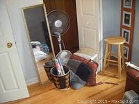 Photo showing large standing mirror with gold frame, floor fan and desk fan on wood floor, woven storage basket, multiple seat cushions and clothes hangers near corner