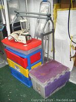 Photo of stacked three-drawer storage unit with bright red, yellow, and blue plastic drawers, three plastic lidded totes in purple and gray, white blender sitting on top, metal clothing rack behind, and small metal step ladder behind the storage units.