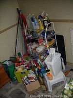 View of the lot showing wire shelving unit filled with various cleaning supplies, brooms, mops leaning against the wall, white plastic waste bins, and assorted boxes and bags on the floor.