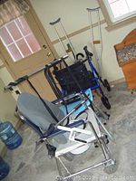 Adjustable shower chair, four-wheeled walker with seat and basket, wheeled commode chair, and pair of underarm crutches seen from doorway in room.