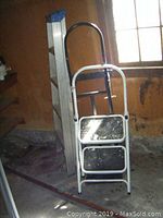 Two step ladders and a hand cart shown grouped together in a corner with window lighting.