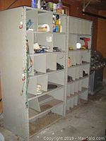 Two large metal shelving units in a garage setting, filled with various tools and supplies including a red cordless drill, yellow work light, bungee cords, and spray bottles. Shelves show wear and surface rust.