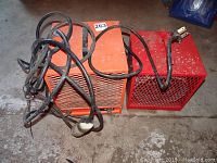 Two industrial electric space heaters lying on concrete floor, one orange and one red, with attached power cords and plugs.