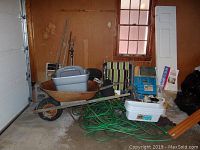 Photo showing a rusty metal wheelbarrow with rubber wheel, coiled green garden hoses on the floor, plastic bins, lawn chairs with striped cushions, and other garden supplies piled in a garage or shed.