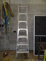 White 5-step step ladder standing upright against a concrete block wall with hanging harnesses on side.