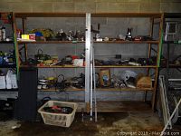Overview of two wooden and metal shelves containing assorted hardware items including tools, clamps, electric motors, belts, wires, and other equipment.