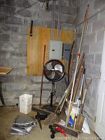 Garage corner showing garden tools arranged against walls with a black standing fan, electrical panels, and a bucket on the floor.