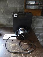 Black and Decker router placed on a wooden surface with a black hard plastic carrying case behind it and power cord coiled in front, showing some sawdust and dirt on the surface.