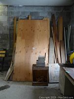 Photo showing large plywood sheets and long wooden planks against a cinder block wall.