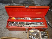 Open red metal toolbox containing various metal hand tools including wrenches and wood spade drill bits with a plastic bag of mixed fasteners on the concrete floor.