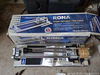RONA tile cutter and box laying on a concrete floor with tools and parts in background.