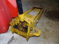 Photo of a yellow heavy duty floor jack with rust and wear marks on a concrete floor next to red gas cans
