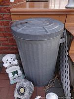 Galvanized garbage can with matching metal lid, white ceramic dog lawn decor holding welcome sign, rock-like solar light, and plastic lattice panel leaning against wall.