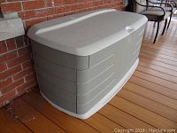 Closed view of Rubbermaid outdoor storage chest resting on wooden deck near brick wall and metal chair.