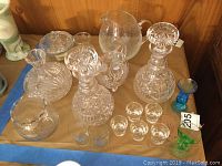 Photo showing multiple cut crystal decanters, clear pitcher, shot glasses, and blue and green cordial glasses on a table.