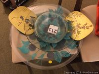 Top view image showing stacked art glass platters with blue and purple leaf and fruit painted design and two yellow wooden Asian style trivets with bird motifs.
