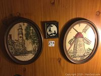 Five fabric art pictures in oval wooden frames showing buildings, a windmill, waterfront, and street scenes, mounted on a wooden wall with a small rectangular silhouette frame in black and white above them.
