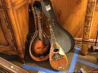 Two vintage pot bellied mandolins with wood bodies standing upright against a wood cabinet, with a single hard case behind them.