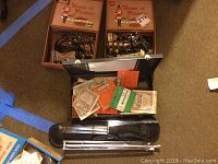 Overhead photo showing open case with multiple boxes of House of Windsor mandolin and guitar strings alongside loose strings and instrument parts on carpeted floor.