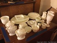 Full view of assorted milk glass items including plates, platters, covered dishes with lids, goblets, and decorative small dishes arranged on a wooden surface.