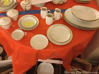 Full view of assorted Noritake Golden Rose dinnerware on orange tablecloth showing plates, bowls, creamers, sugar bowls, and teacups with saucers arranged.