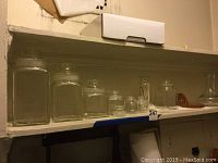 Shelving with 6 glass apothecary jars with lids, arranged by size left to right.