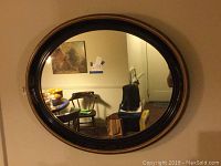 Oval black framed wall mirror with gold accents, approximately 32x26 inches, hanging on a beige wall reflecting a dining area