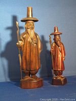 Two hand carved wooden figures of robed men holding staffs, standing on octagonal bases against blue background.