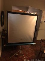 Full view of the Colormaster Radiant collapsible movie screen fully extended on tripod stand inside a room with wooden flooring.