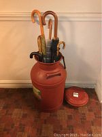 Full view of the metal milk can holding six umbrellas inside, shows can's reddish-brown color and umbrella handles