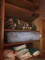 Photo of brown plaid blanket, blue floral embroidered blanket, and assorted folded linens on shelves