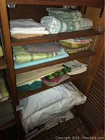 Wooden shelving unit with multiple folded linens including white and green patterned table cloths and pillows on lower shelves