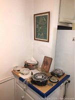 Photo showing framed cross-stitch sampler, glass bowls, trivet, heating warmer, ashtray, and additional decor items on a counter beneath a framed blessing wall art.
