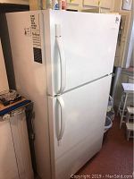 Full view of white top mount refrigerator freezer with freezer on top and refrigerator below, visible handles, placed in a room.