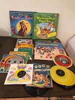 Photo shows multiple children's records including Lady and The Tramp, Winnie the Pooh and Tigger Too, Pinocchio, with visible covers and colorful illustrations. Yellow colored discs are visible along with black vinyl records.