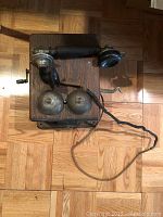 Full view of the antique wooden wall-mounted telephone showing dual metal bells, black handset, and crank handle on hardwood floor.