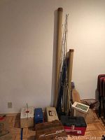 Overall view of fishing rods propped against wall, multiple tackle boxes on floor, wooden filet board and fishing net visible.