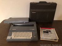 Smith Corona gray Model SC 104 typewriter with white keyboard, detachable cover placed on top, and black carrying case in background on brown surface