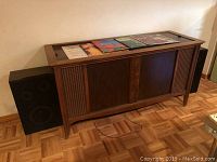 Side view of vintage Magnavox wooden record cabinet with vinyl records on top and one speaker beside it.