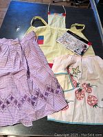 Photo of three items on a table: yellow apron with patch pockets, pink embroidered apron, and a vintage sewing pattern booklet.